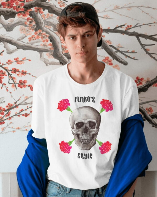 Modelo Chico joven con una gorra negra hacia atrás posa frente a un fondo decorativo de ramas de árbol y flores. Lleva puesta una camiseta blanca con un diseño en el centro que muestra una calavera rodeada de cuatro claveles rojos. El texto "Flmko's Style" aparece tanto en la parte superior como en la inferior de la calavera. El joven lleva una chaqueta azul que descansa sobre sus hombros, creando un contraste interesante con el fondo floral y el diseño oscuro de la camiseta.