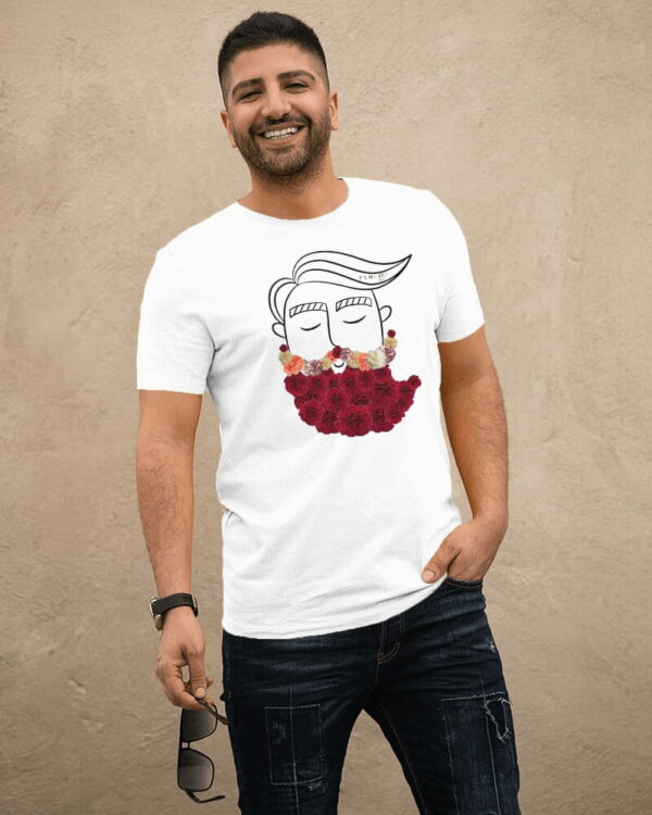 Un hombre sonriente está posando mientras usa una camiseta blanca con un diseño artístico en la parte delantera. El diseño minimalista en la camiseta muestra un dibujo de un rostro con una barba compuesta por flores de colores, combinando simplicidad con un toque creativo. El hombre lleva unos jeans oscuros y sostiene un par de gafas de sol en su mano izquierda, mientras posa relajado y natural contra un fondo neutro.